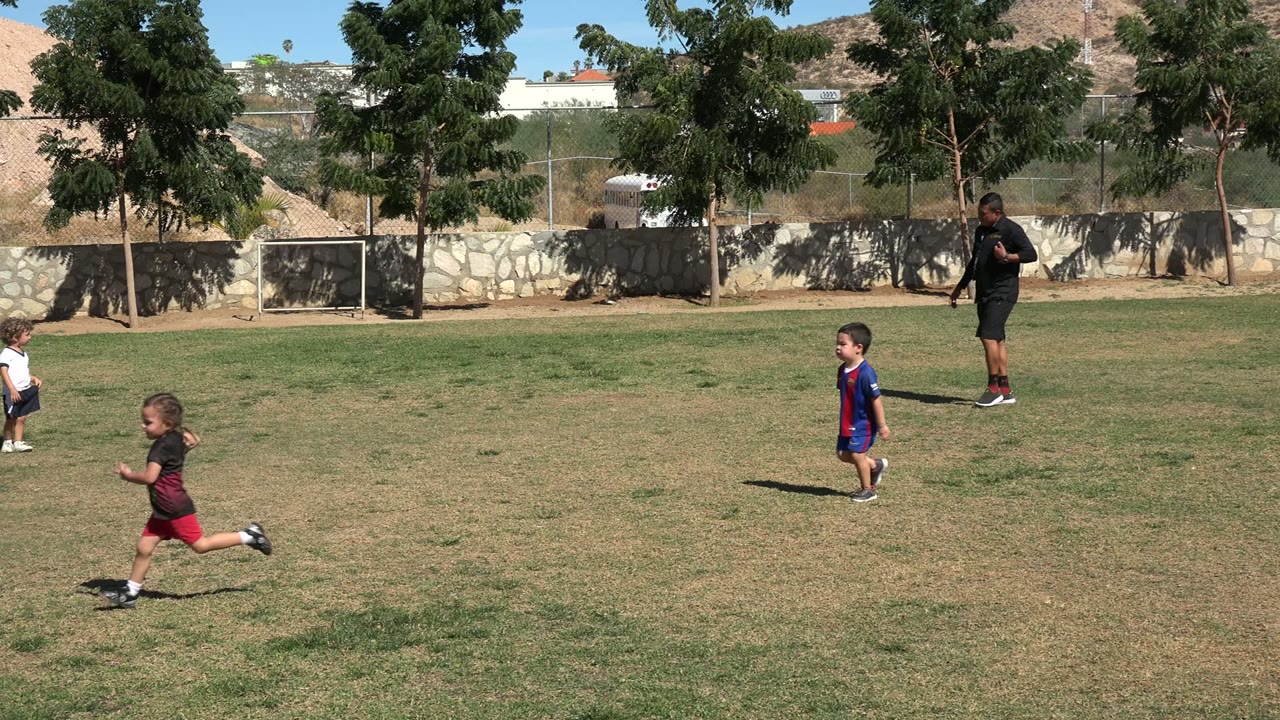A child in red shorts runs across the midground in Los Cabos as another child and an adult remain near the trees behind the field.