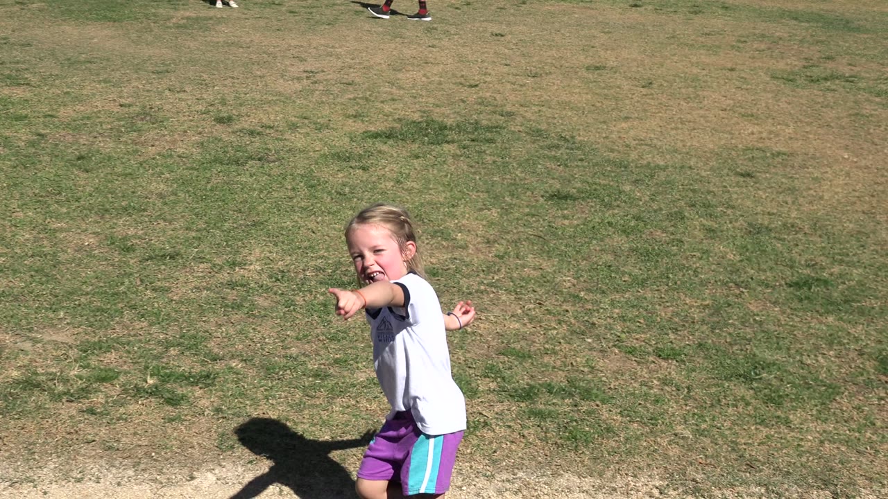 Blonde girl in a white soccer shirt points while standing near a grassy field.