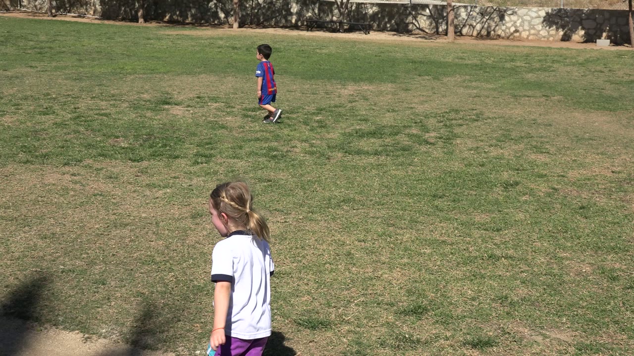 The girl is seen from behind on the patchy grass field in Los Cabos, Mexico, with another child in a striped soccer jersey farther upfield.