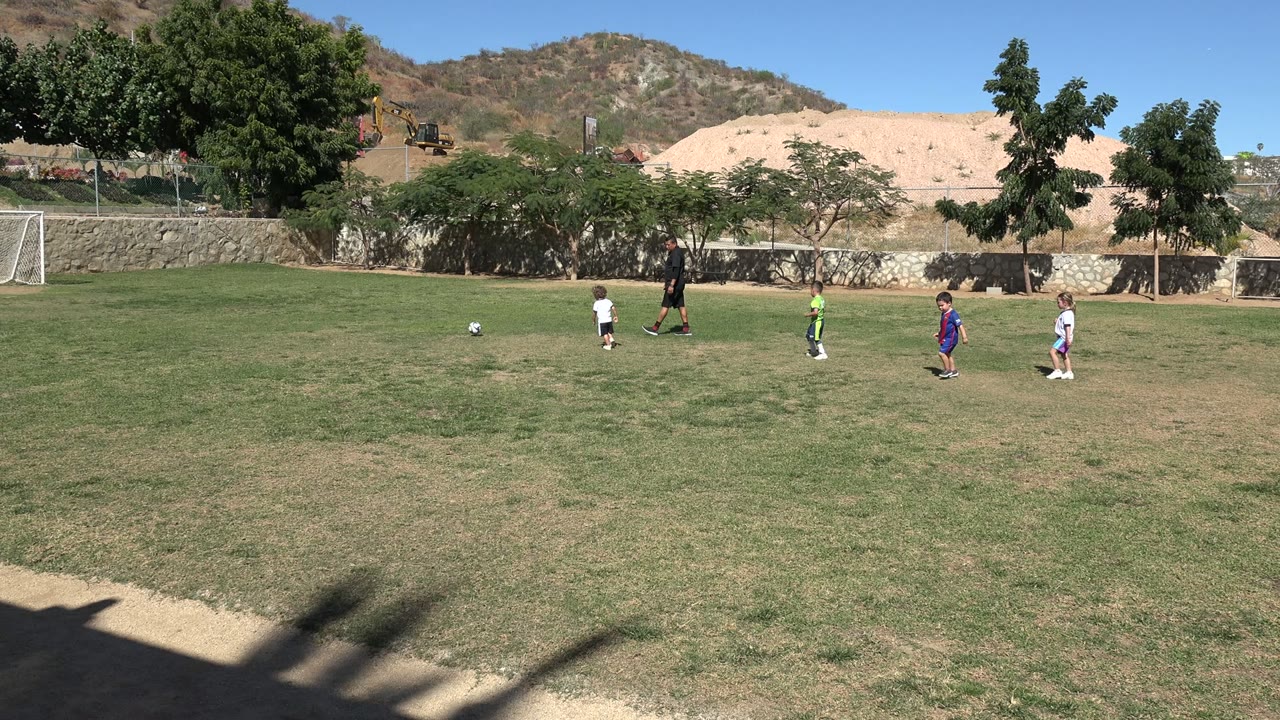 Wide view of children and an adult on a grass soccer field in Los Cabos, Mexico, with white goals, trees, a stone wall, tan earth, and hills in the background.