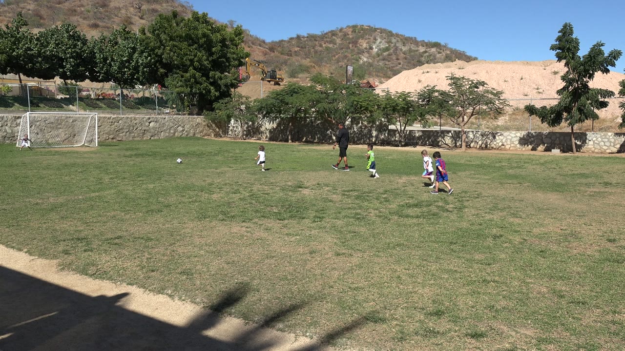 A wide Los Cabos field frame shows children clustered toward the right, an adult near the center, a soccer ball on the left, and a goal set against trees and hillside.