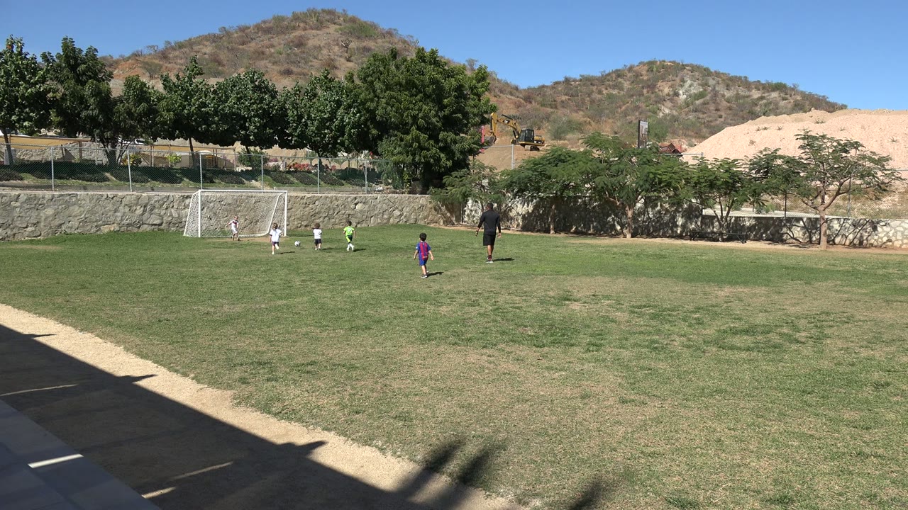 Children in light and green jerseys gather near the white goal on the Los Cabos soccer field while another child in a purple top stands farther out on the grass.