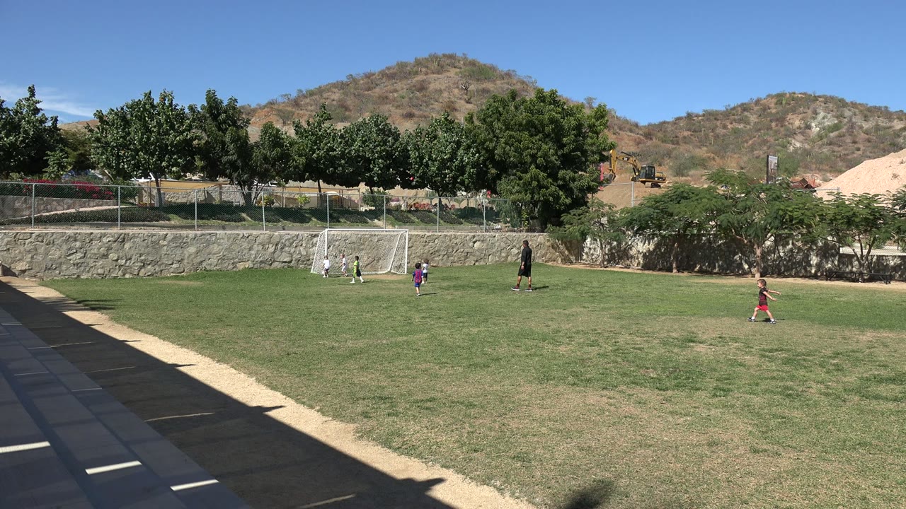 Children cluster closer to the small soccer goal while the teacher stands to the right on the sunny Los Cabos field, with trees, fencing, and hills behind them.