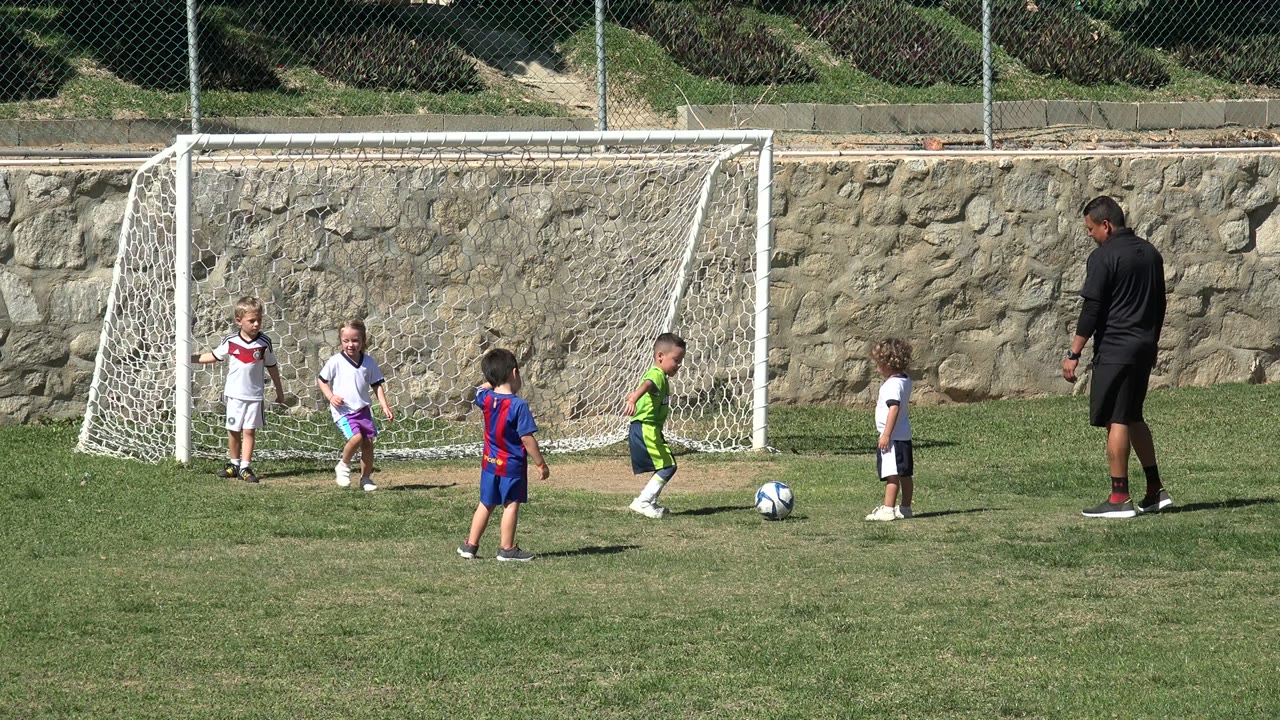 Children in white, green, and striped jerseys stand near a soccer ball in front of a white goal in Los Cabos, Mexico, with the adult coach to the right.