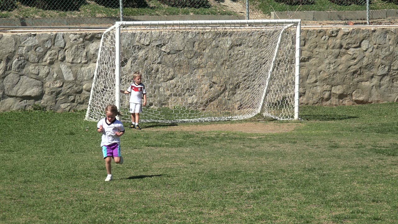 The running child is nearer the center of the Los Cabos soccer field, while another child remains near the white goal in the background.