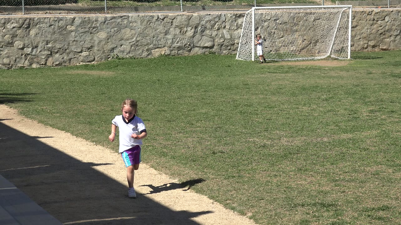 At the start of the Los Cabos clip, a young girl runs along a sidewalk beside grass, with a small soccer goal and stone wall in the background.