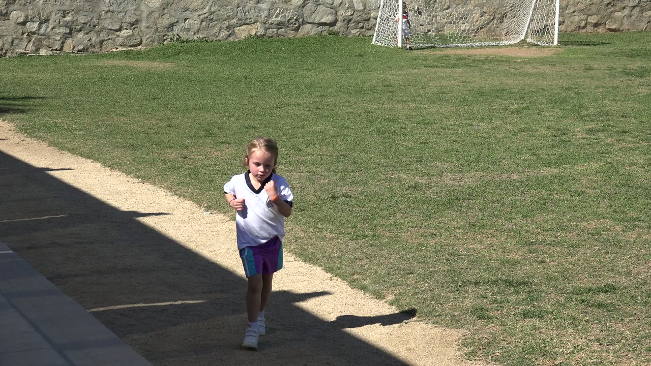 One second in, the girl is closer to the camera on the sidewalk, with sunlit grass, a small goal, and a long shadow line behind her in Los Cabos.