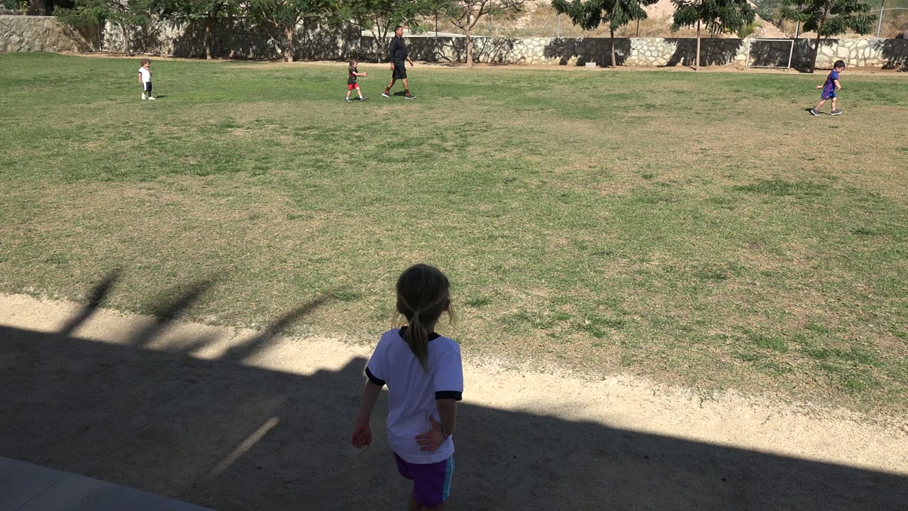 The child faces away from camera in the shaded sideline area in Los Cabos as children and an adult remain spread across the bright grass field.