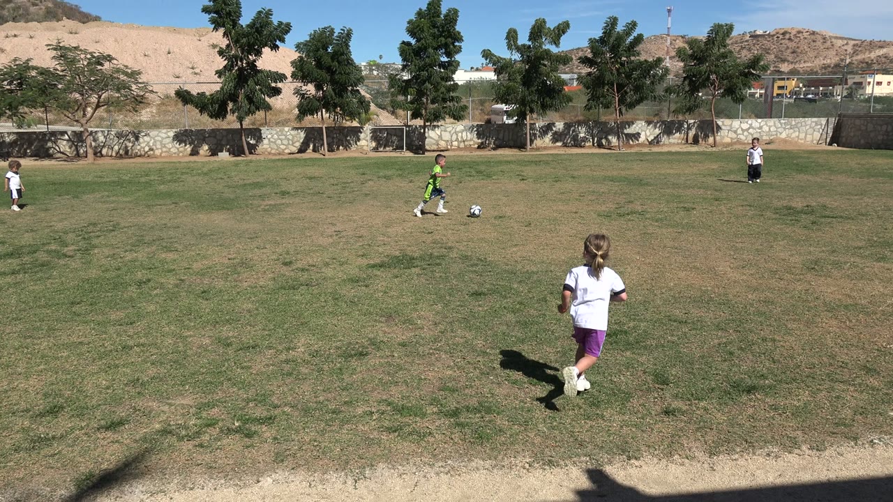 A child in a green soccer jersey moves near a soccer ball on a patchy grass field in Los Cabos, Mexico, with another child in white farther back.