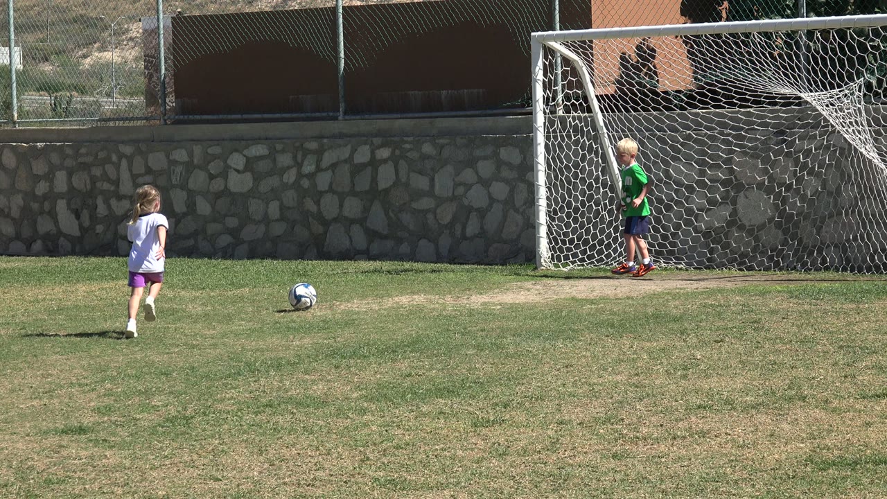 In Los Cabos, Mexico, a girl in a white shirt and purple shorts runs toward a soccer ball while a blond child stands inside the white goal.