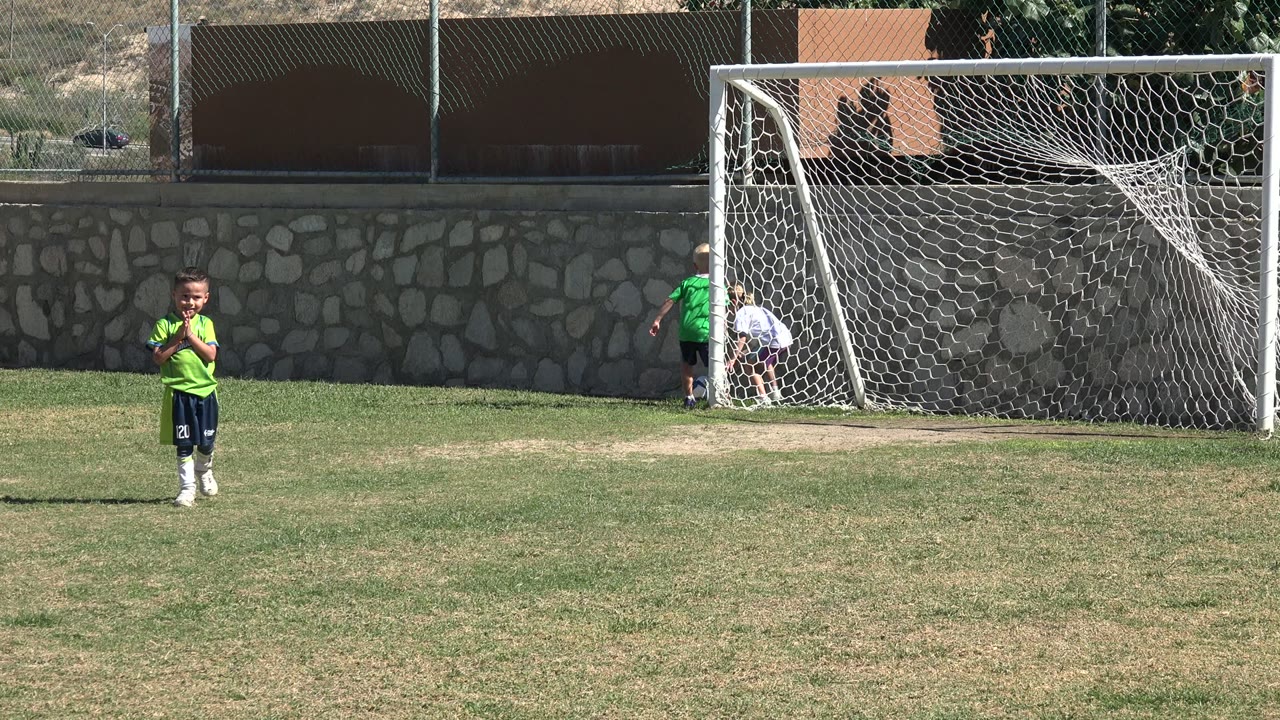 Child in green jersey on a grass field near a white soccer goal.