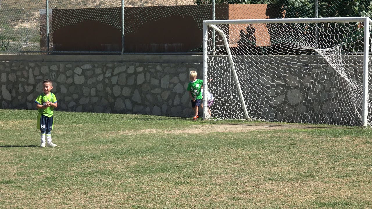 At 0:01 in Los Cabos, Mexico, the foreground child faces the camera on the grass as children remain close to the white net and stone wall in the background.