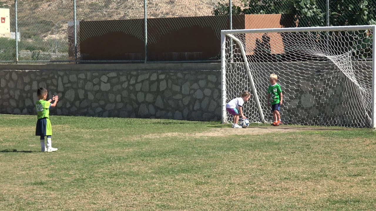 A child in a white shirt bends over the soccer ball near the goal while another child in a green jersey stands inside the net area in Los Cabos, Mexico.