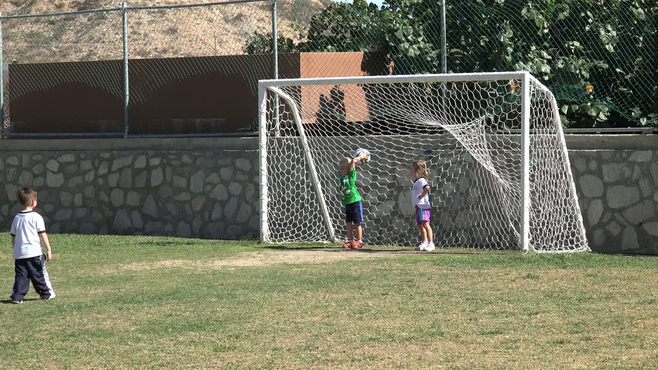 Two children remain inside the white goal in Los Cabos, Mexico, while another child stands in the foreground facing the net.