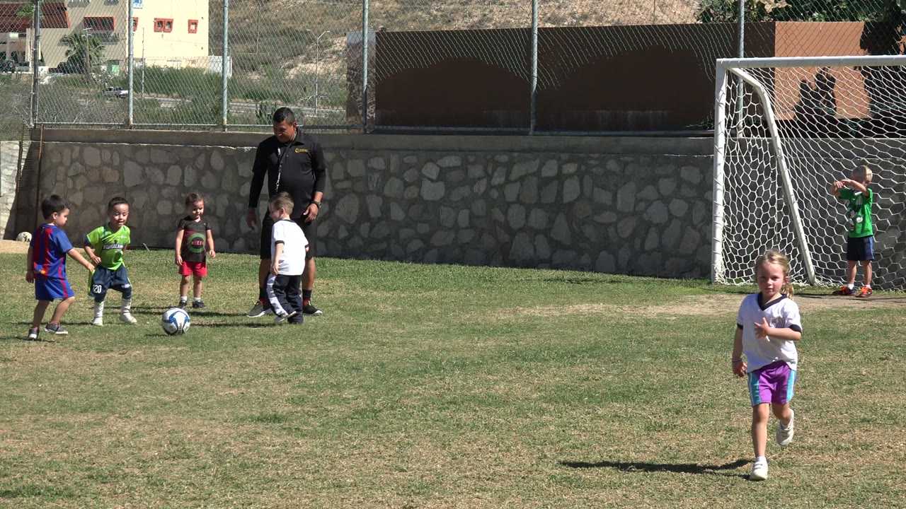 Several small children in bright soccer shirts cluster around the ball on a Los Cabos field, with an adult coach watching and a white goal partly visible to the right.