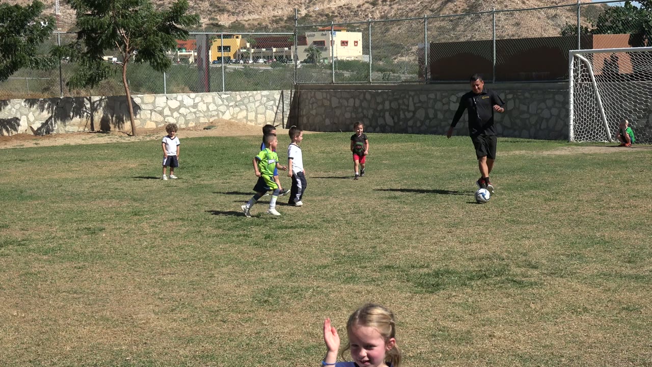 Young soccer players stand spaced out on the grass in Los Cabos, Mexico, as the coach remains near the ball with the goal net in the background.