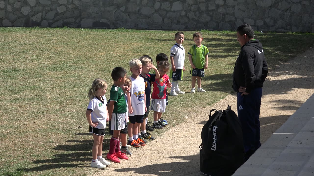 Children in soccer uniforms stand in a line during practice in Los Cabos, Mexico, with a coach at right and a black equipment bag in the foreground.
