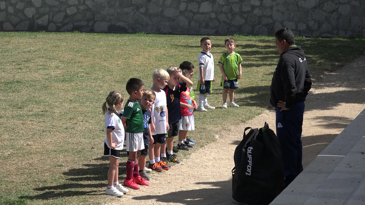 A line of young soccer players remains on the grass in Los Cabos, Mexico, with two children set farther back near the stone wall and the coach standing at right.