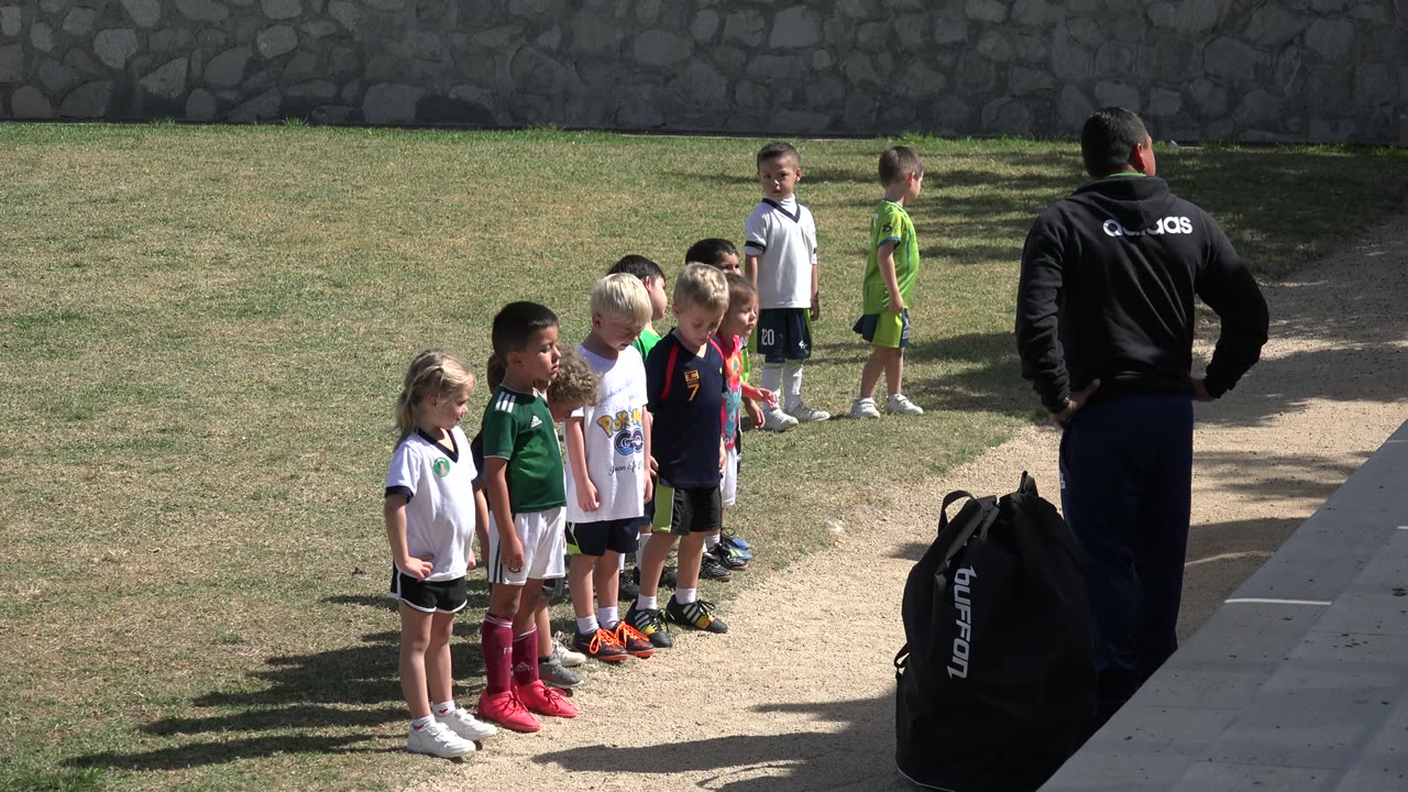 Children lined up outdoors as a man in a black jacket watches nearby.
