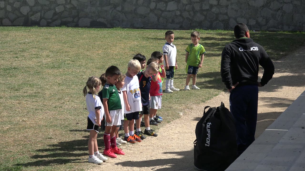 A line of children faces toward an adult in a black jacket in Los Cabos, Mexico, with the group lit by direct sun and a stone wall behind them.