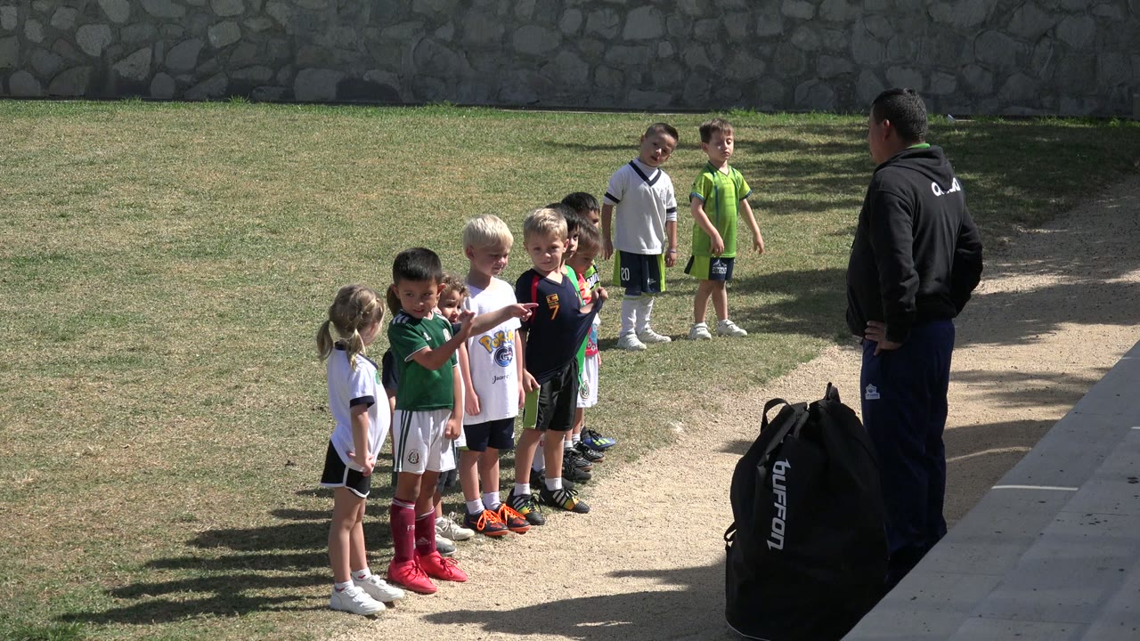 One second into the Los Cabos clip, the children remain lined up on the field while the coach stands at the right edge near the equipment bag.