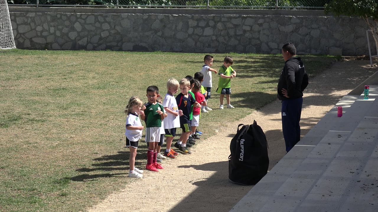 The group of children shifts closer together on the Los Cabos field as the adult instructor watches from the paved edge beside the sports bag.