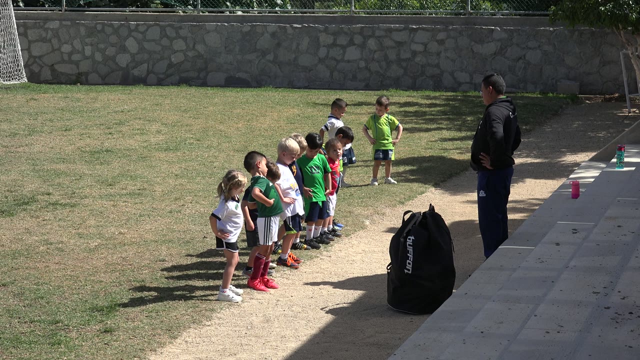 Young children stand in a line on a grass field in Los Cabos, Mexico, while an adult coach faces them near a black sports bag and paved sideline.