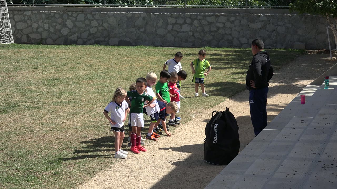 The children remain lined up on the Los Cabos grass field, with several looking down or sideways as the adult coach stands near the paved edge and sports bag.