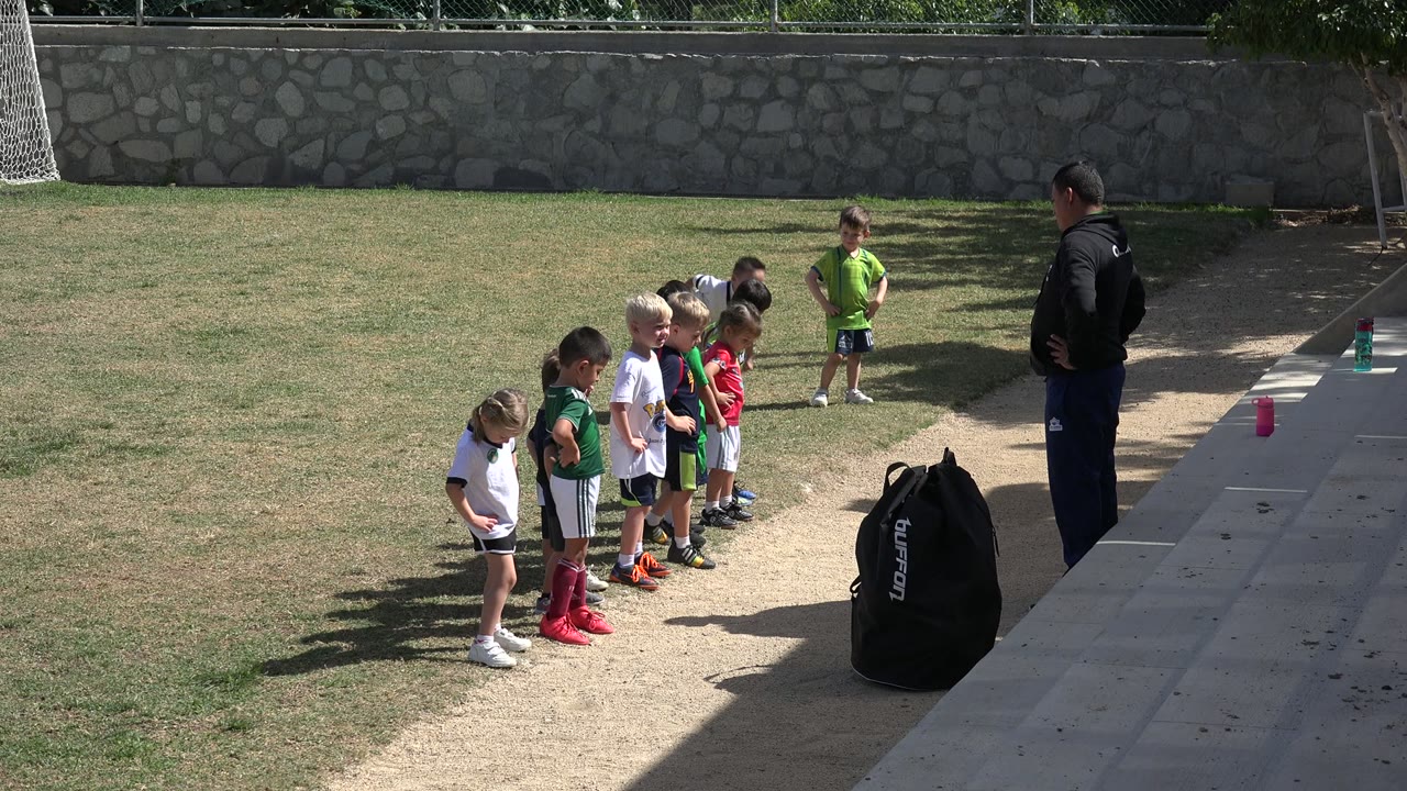 Children in soccer uniforms stand in a sideline line at a Los Cabos, Mexico soccer practice while a coach faces them near a paved edge.