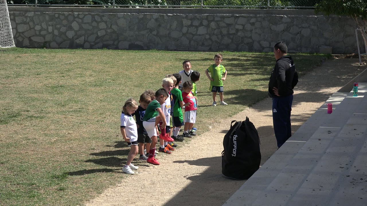 The Los Cabos youth soccer group remains lined up on the field, with several children facing the coach and bright sunlight cutting across the grass.