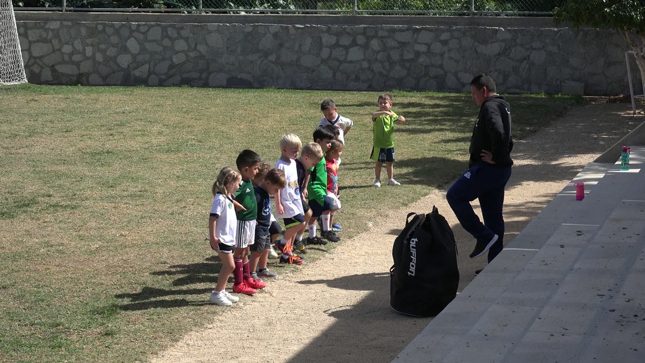 At 0:00 in Los Cabos, Mexico, young children in white and green uniforms line up on a grass field while an adult instructor stands near concrete steps.