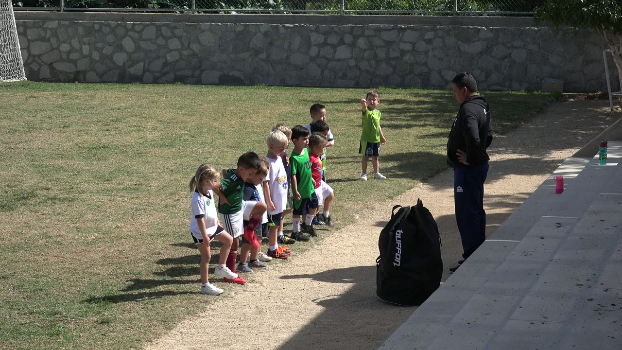 At 0:01 in Los Cabos, Mexico, several children lean forward in an exercise drill as the instructor stands to the right near a black gear bag and concrete steps.