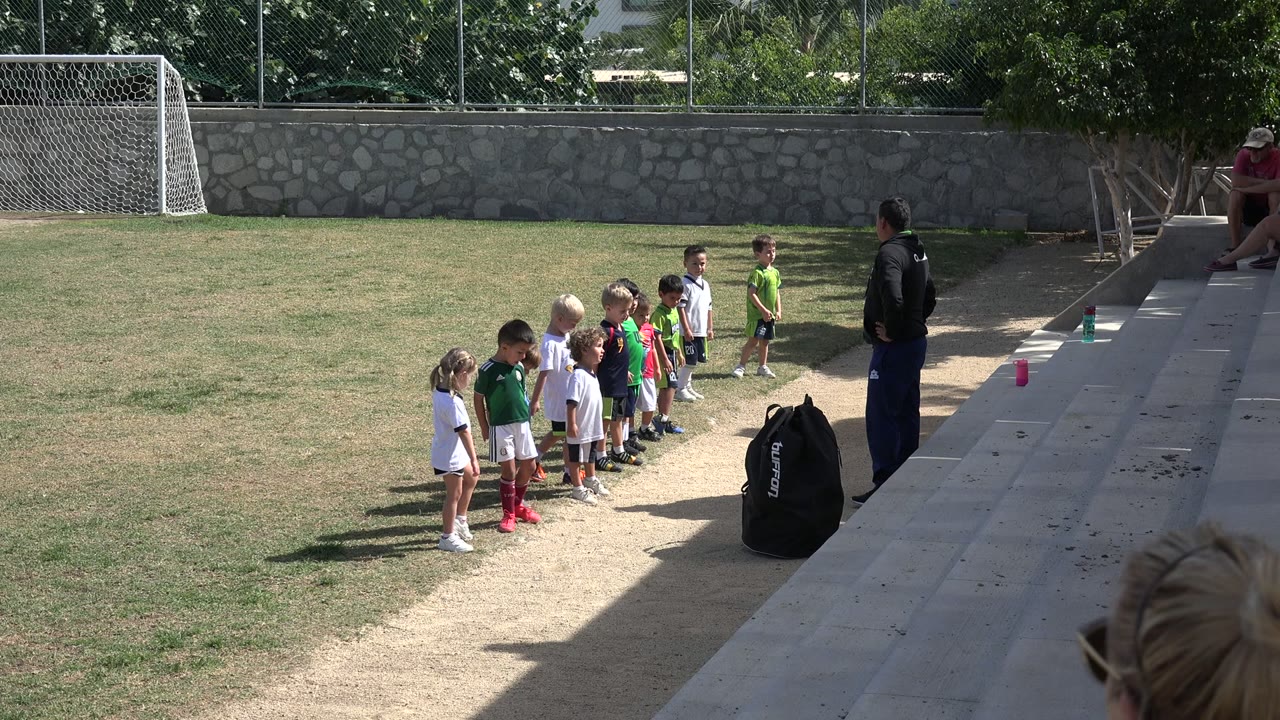 Small children stand in a line on a Los Cabos soccer field while an adult coach faces them near concrete steps and a black gear bag.