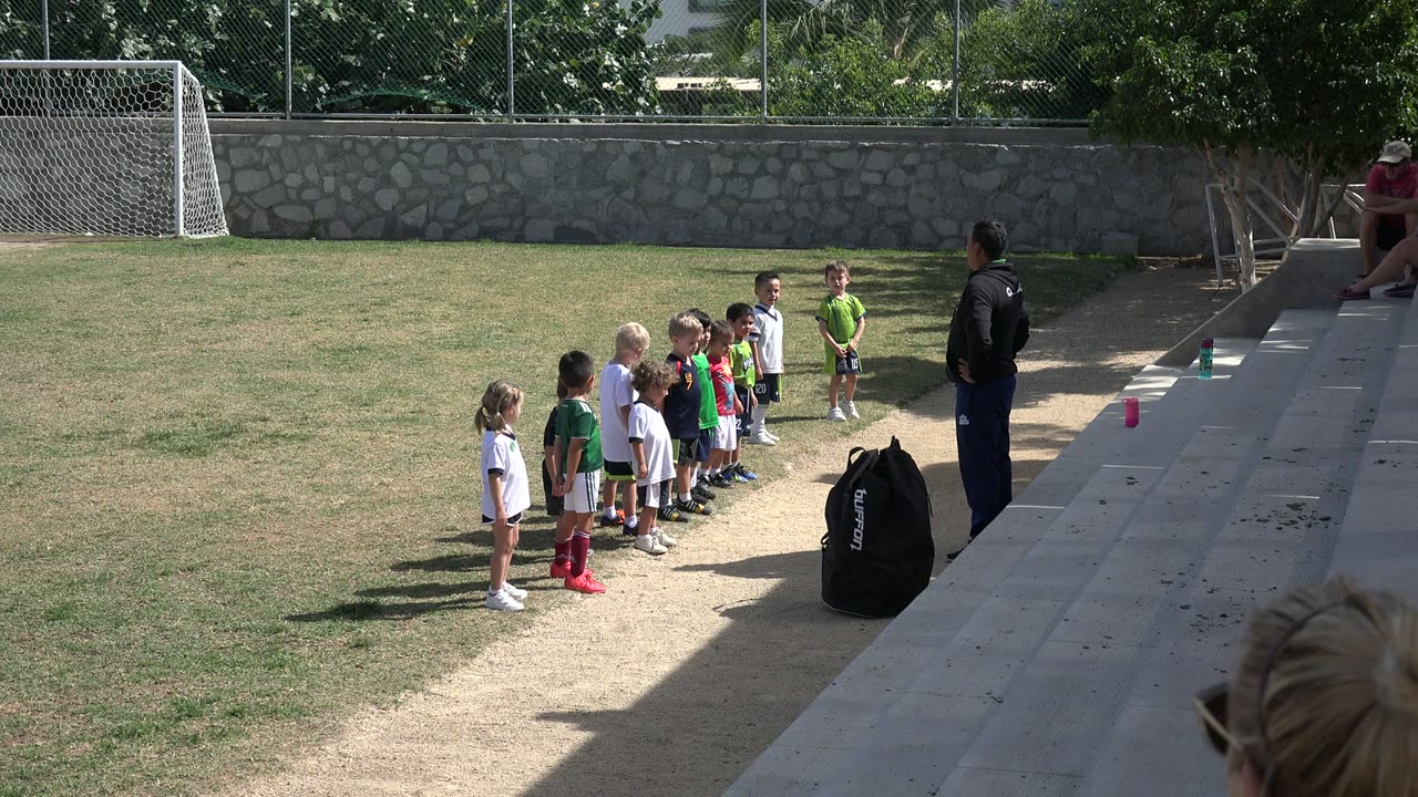 The line of children remains near the sideline in Los Cabos, with the coach standing by the steps and the soccer goal set against a stone wall behind them.