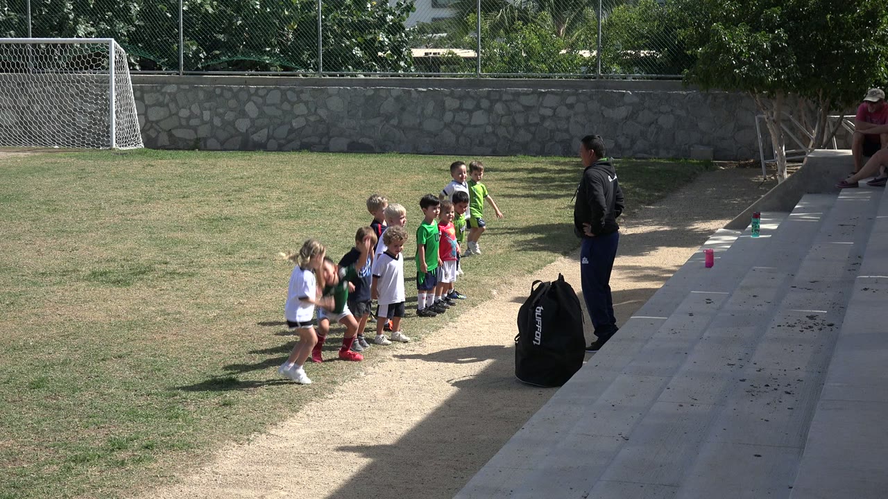 In Los Cabos, Mexico, the children start an exercise movement on the grass while the instructor stands at the edge of the dirt path near a black sports bag.