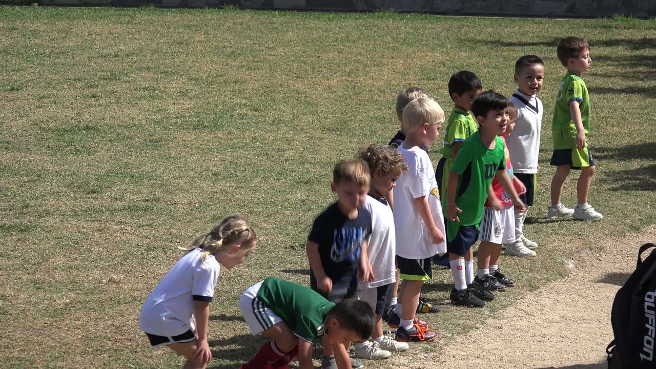 Several children crouch or lean forward while others stand behind them on a sunlit grass field in Los Cabos, Mexico, with green team shirts prominent in the group.