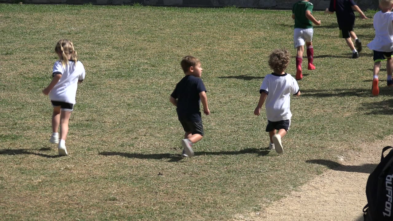 The children continue running across the Los Cabos field, with several kids in white and dark shirts moving toward the upper right and strong shadows on the grass.