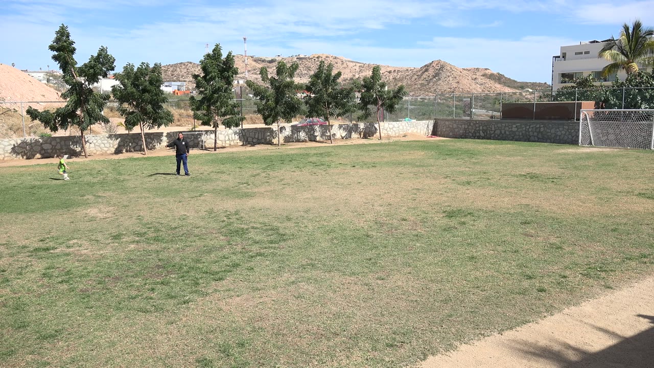 Wide frame in Los Cabos, Mexico showing children on the left edge and an adult standing on a sunlit grass field with trees, fencing, hills, and buildings in the background.