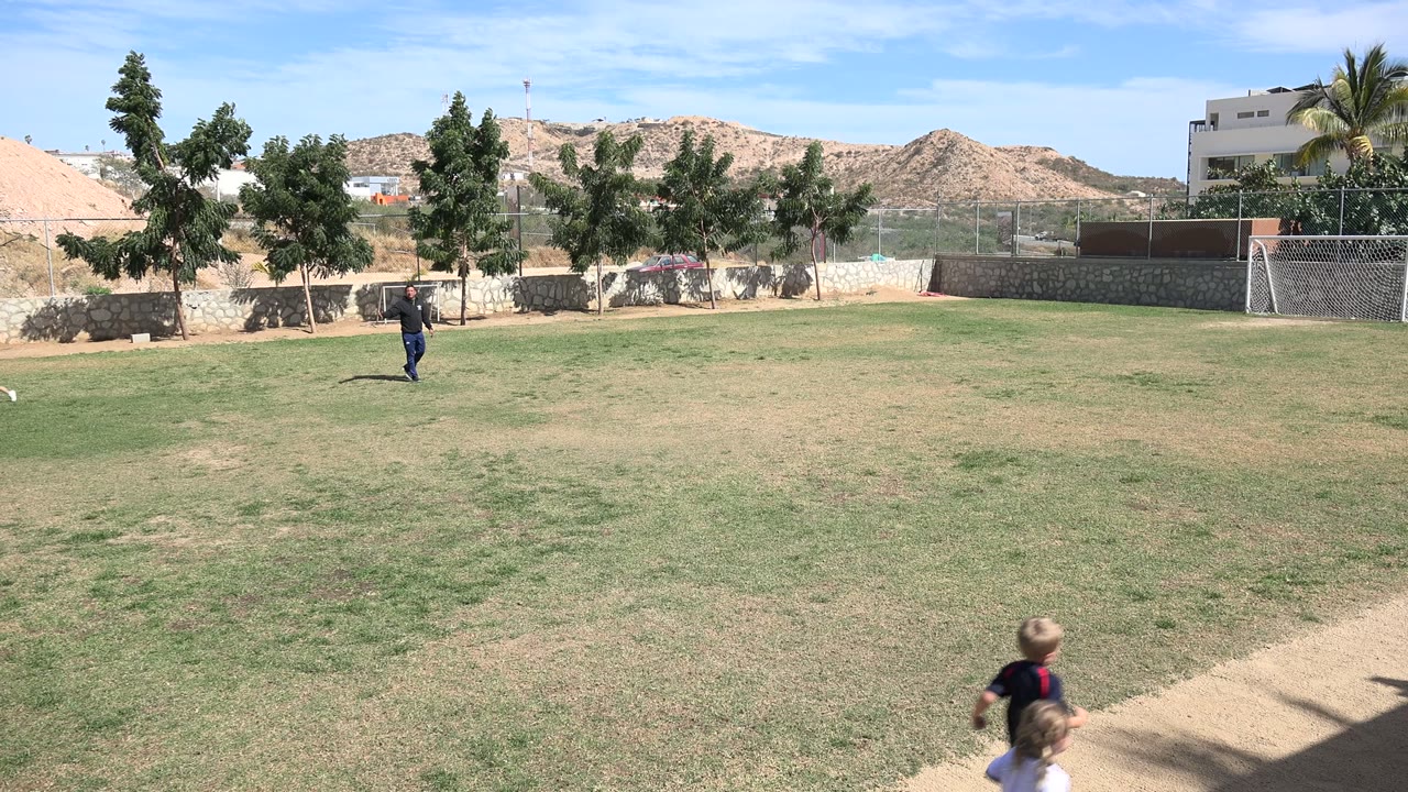 Wide Los Cabos soccer-field view with an adult in dark clothing near the midground and two children partly visible at the lower right foreground.