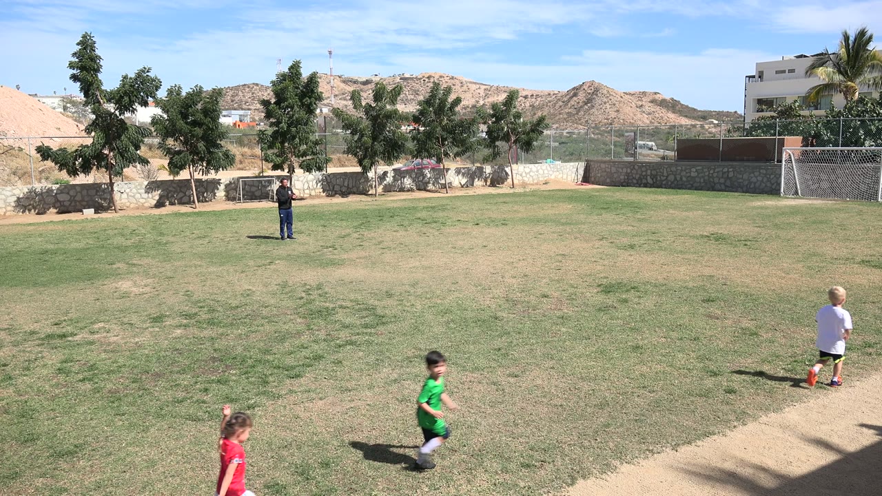 Children running on a grass football field with an adult in the distance.