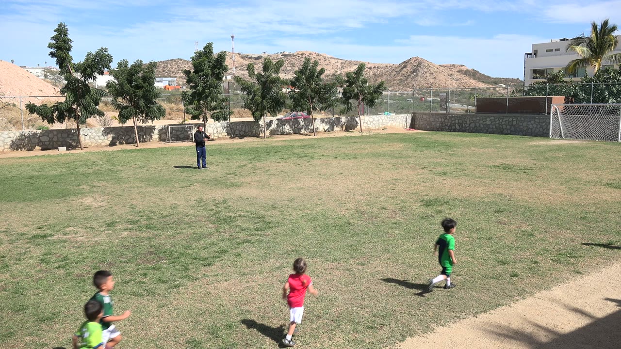 At 0:01 in Los Cabos, Mexico, children in red, green, and white shirts move rightward across the field, with a standing adult still visible near the far sideline.