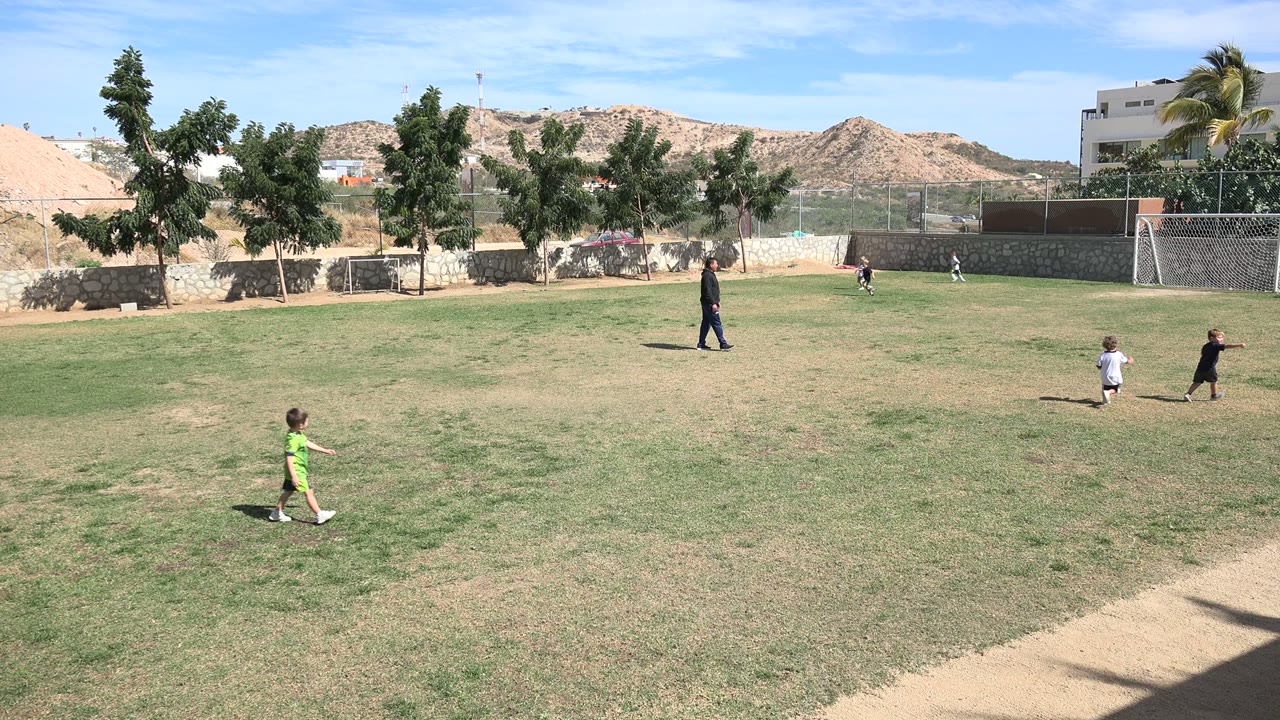 Wide Los Cabos field view with the child in green moving across the left foreground, two children running near the center-right, the coach standing mid-field, and hills and buildings behind the trees.