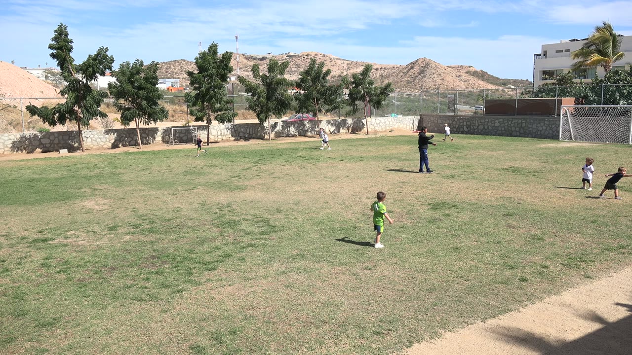 Wide frame of children playing football on a grassy field in Los Cabos, Mexico, with an adult standing mid-field, small goals, trees, walls, buildings, and dry hills in the background.