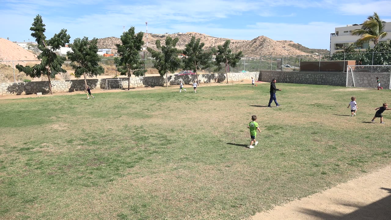 Children continue playing football across the open grass in Los Cabos, Mexico, with one child in a green shirt in the foreground and small goals and hillside buildings behind the play.