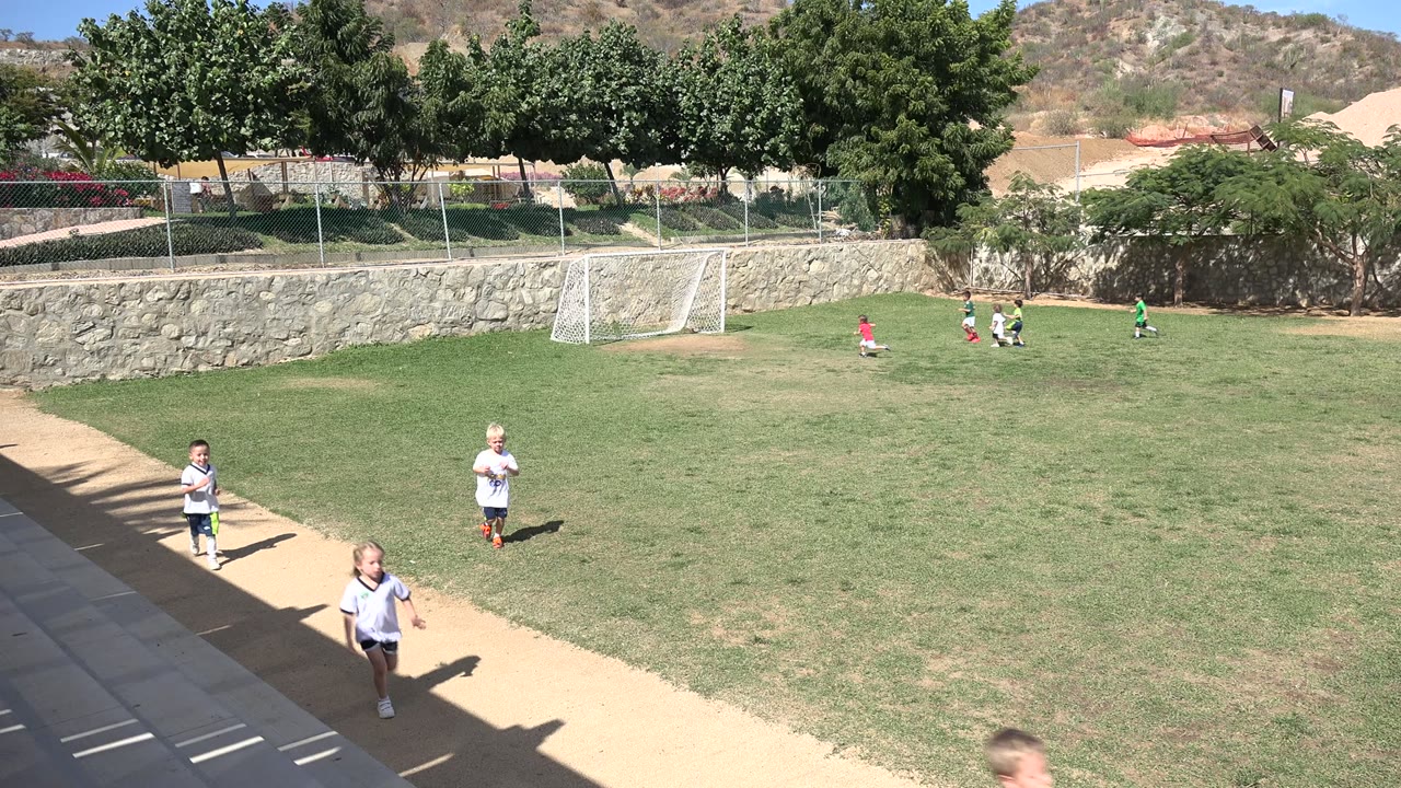 Wide view of children playing soccer on a Los Cabos field, with two children near the foreground walkway and a small goal in the distance.