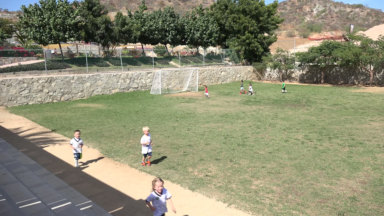 Two young soccer players run closer to the camera on the walkway edge in Los Cabos, while several children cluster near the goal in the background.