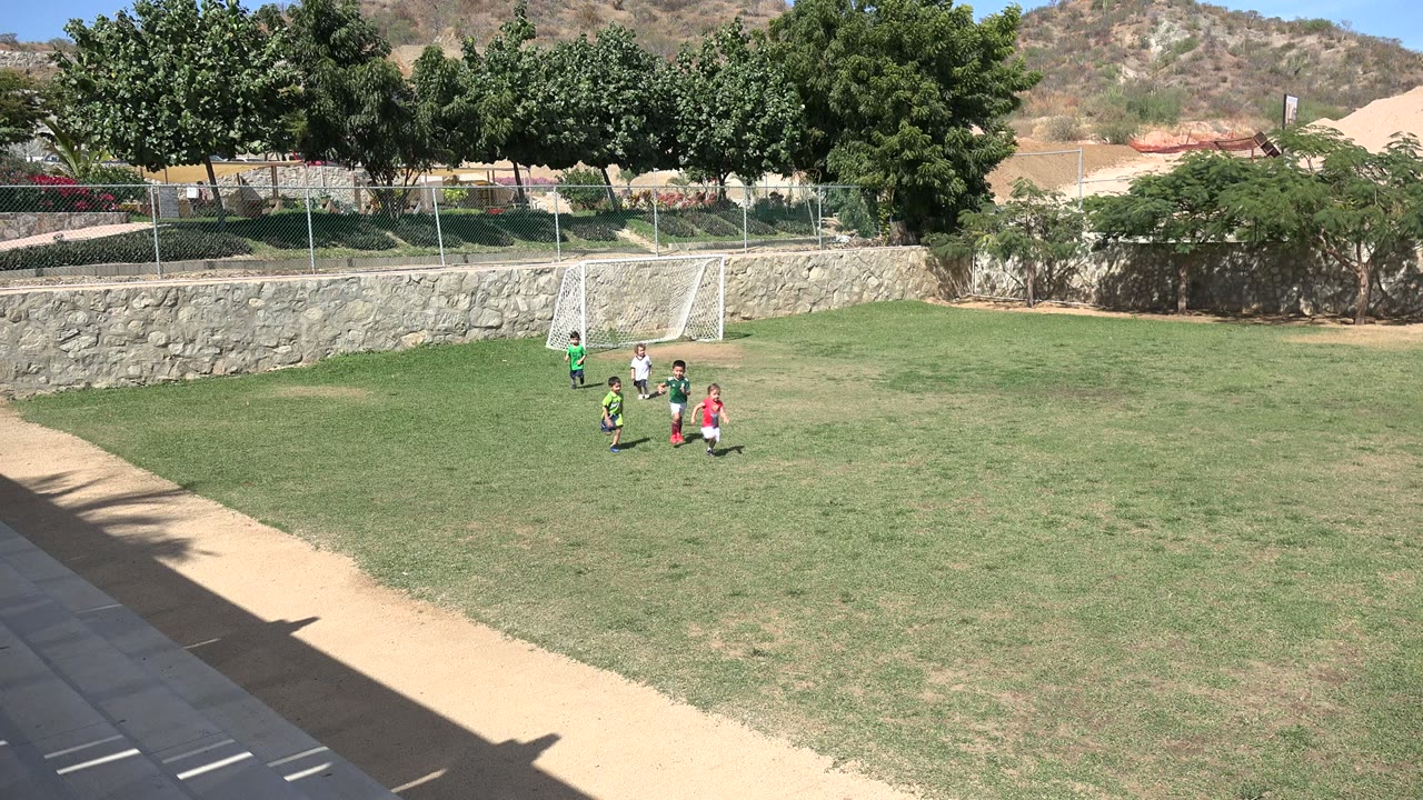 Children in green, white, dark, and red shirts run across the soccer field in Los Cabos, Mexico, with the white goal set behind them.