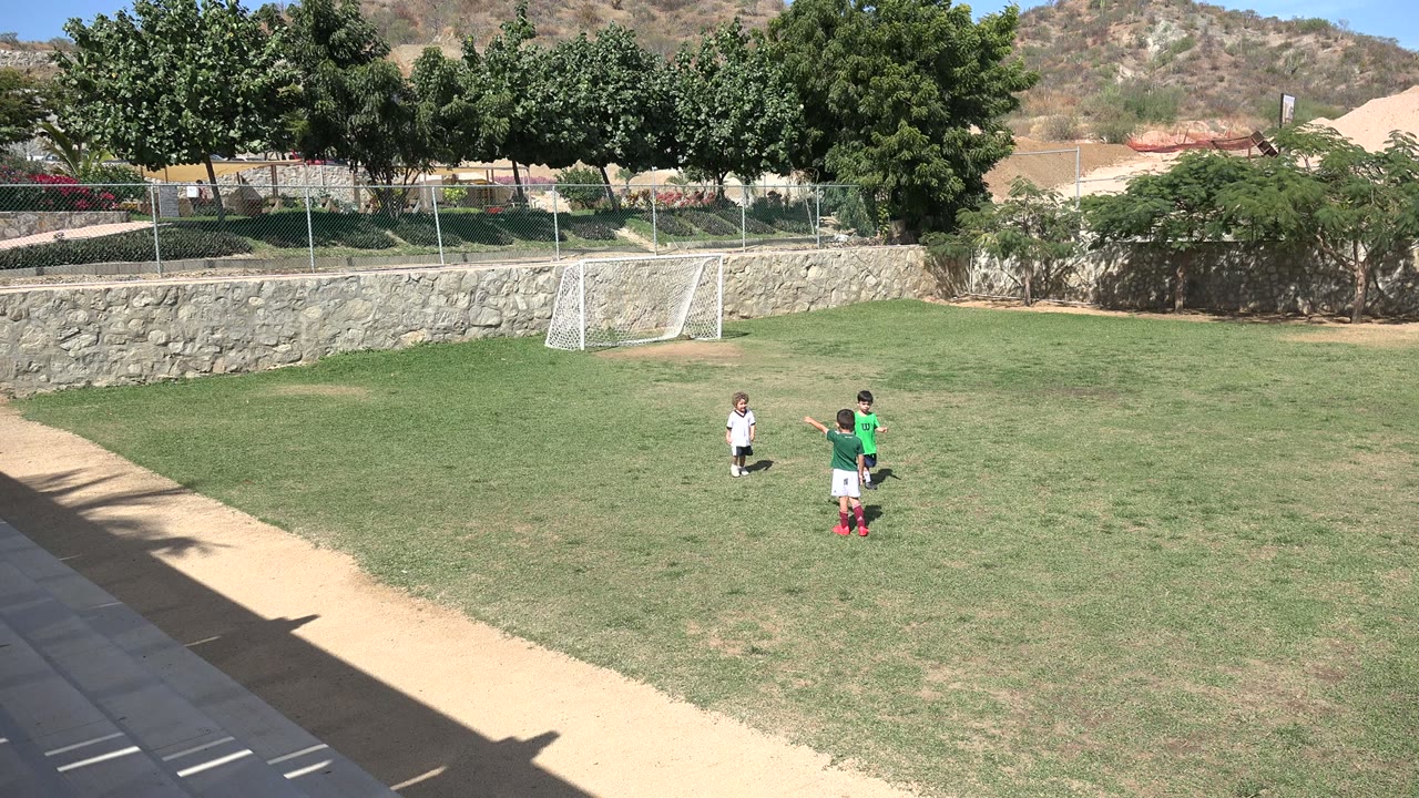 The children start to separate on the Los Cabos soccer field, with one child in a green jersey beside another child and the small goal centered behind them.