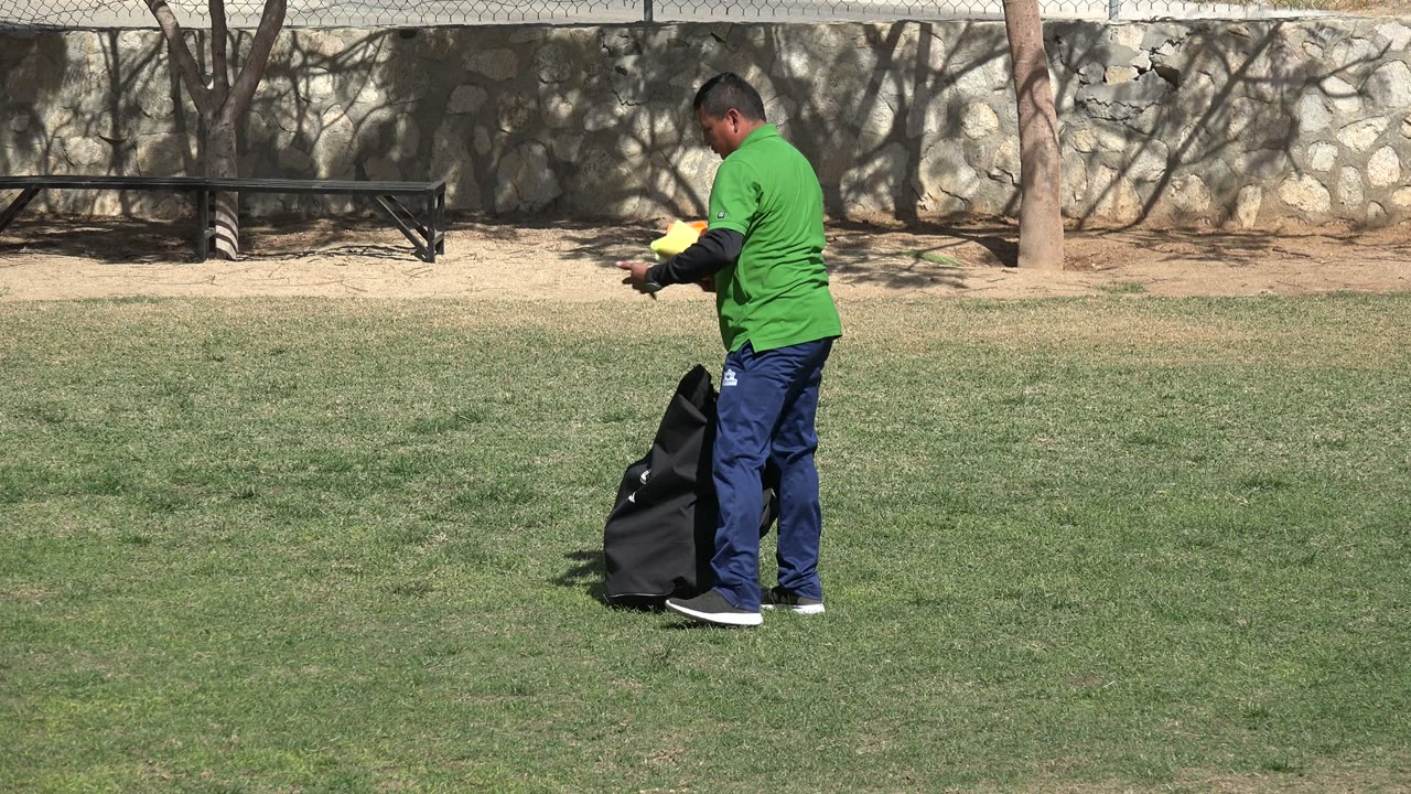 In Los Cabos, Mexico, the man stands next to the open black bag while holding bright cones over the grass, with the wall and fence in the background.
