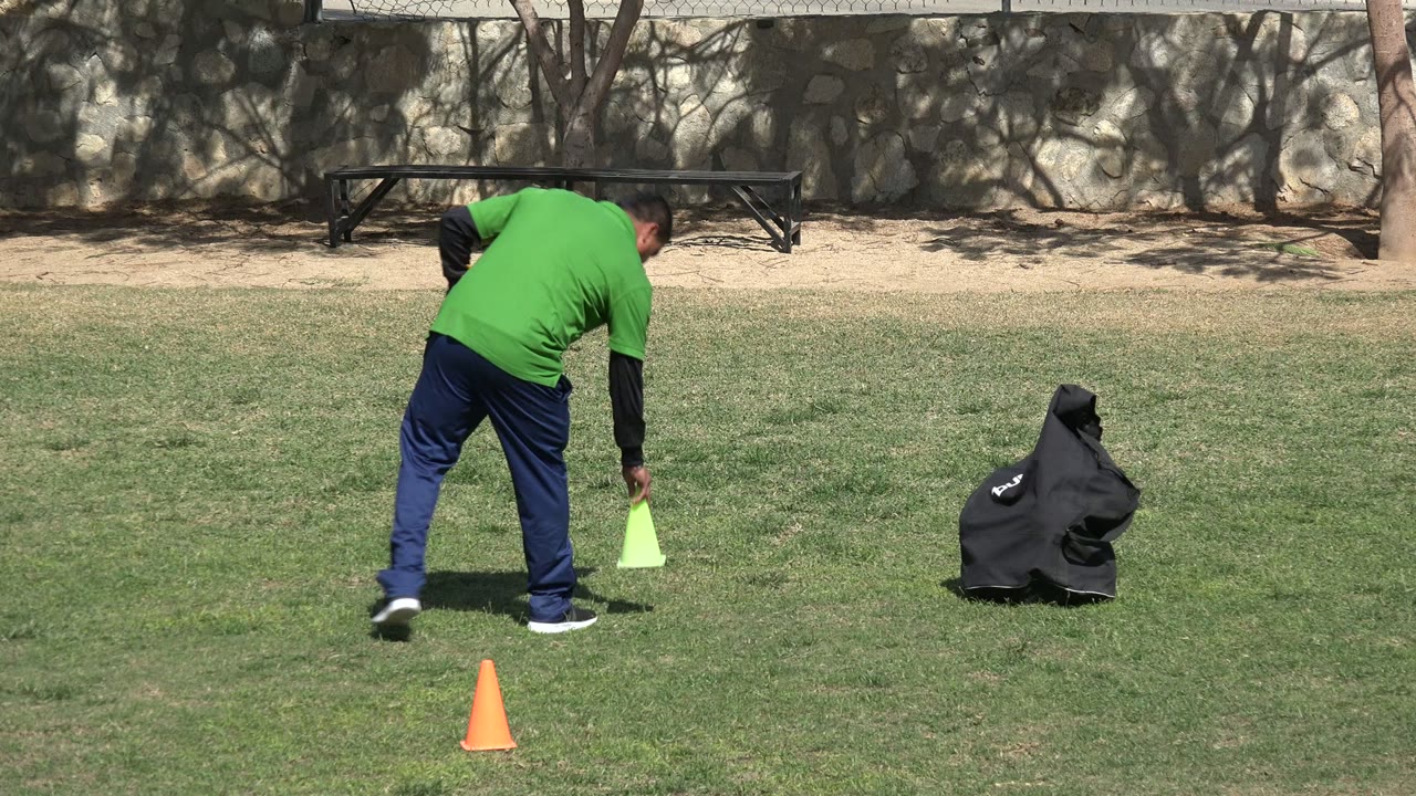 In Los Cabos, Mexico, the coach bends forward to set a lime training cone on the grass while an orange cone and black equipment bag remain nearby.